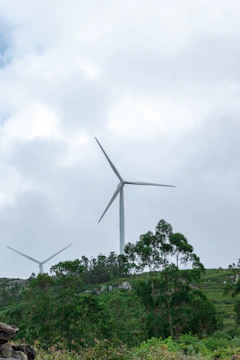 Tall white wind turbines spinning gracefully on a green hillside against a bright, partly cloudy sky.