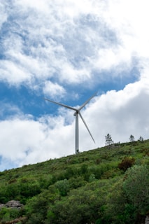 A large wind turbine stands prominently against a backdrop of partly cloudy skies. It is situated on a hill covered with lush green vegetation and some trees.