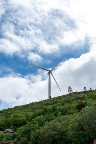 A large wind turbine stands prominently against a backdrop of partly cloudy skies. It is situated on a hill covered with lush green vegetation and some trees.
