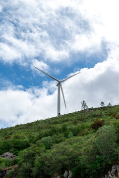 A wind turbine stands tall on a hill covered in lush green vegetation with a partly cloudy sky in the background.