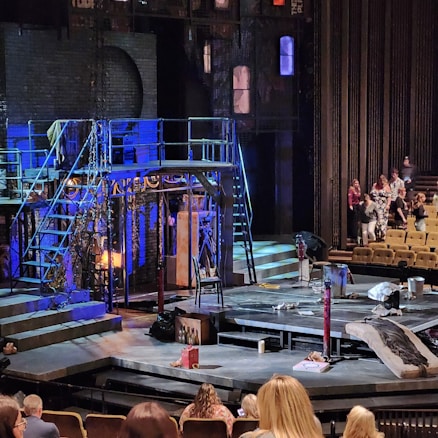 A theater stage set up with various props, including scaffolding, chains, and lighting equipment. The backdrop mimics a gritty urban environment. Several people are seated in the audience, and a few are standing, suggesting either a break or preparation before a performance begins.
