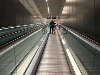 A long, indoor moving walkway with reflective glass walls on either side, leading to an illuminated end with signs hanging from the ceiling. Several people are visible in the distance, walking and pushing bicycles.
