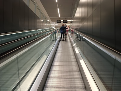 A long, indoor moving walkway with reflective glass walls on either side, leading to an illuminated end with signs hanging from the ceiling. Several people are visible in the distance, walking and pushing bicycles.