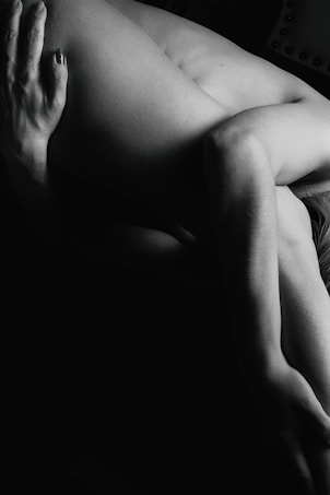 A black and white photograph features a person in an artistic pose, highlighting the contours and shadows of the human form. The lighting creates a dramatic effect, focusing on the texture and shape of the limbs.