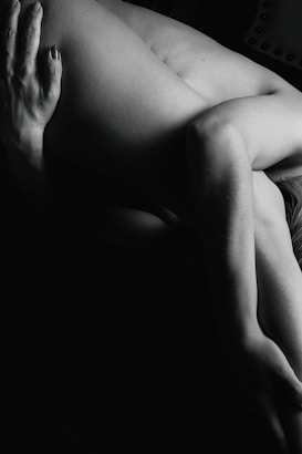 A black and white photograph features a person in an artistic pose, highlighting the contours and shadows of the human form. The lighting creates a dramatic effect, focusing on the texture and shape of the limbs.