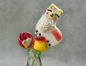 A whimsical arrangement of a llama-shaped confection, decorated with white icing and small pink accents, placed alongside colorful candies. The sweets are displayed in a glass bottle, creating a playful combination of desserts.