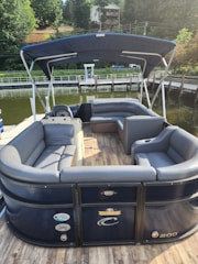 A pontoon boat with sleek gray and blue seating is moored by a wooden dock. The boat features a blue canopy providing shade over the seating area. The background includes a body of water, a dock, and a hillside with lush trees and a house.