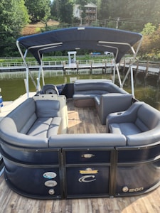 A pontoon boat with sleek gray and blue seating is moored by a wooden dock. The boat features a blue canopy providing shade over the seating area. The background includes a body of water, a dock, and a hillside with lush trees and a house.