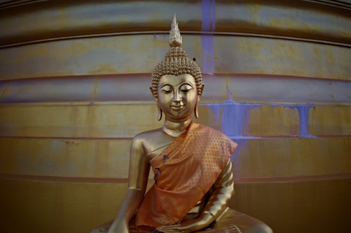 A golden statue of the Buddha sitting in a meditative posture. The statue is adorned with an intricate orange robe with detailed patterns. It is set against a large, smooth backdrop with golden hues and traces of blue stains.