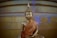 A golden statue of the Buddha sitting in a meditative posture. The statue is adorned with an intricate orange robe with detailed patterns. It is set against a large, smooth backdrop with golden hues and traces of blue stains.