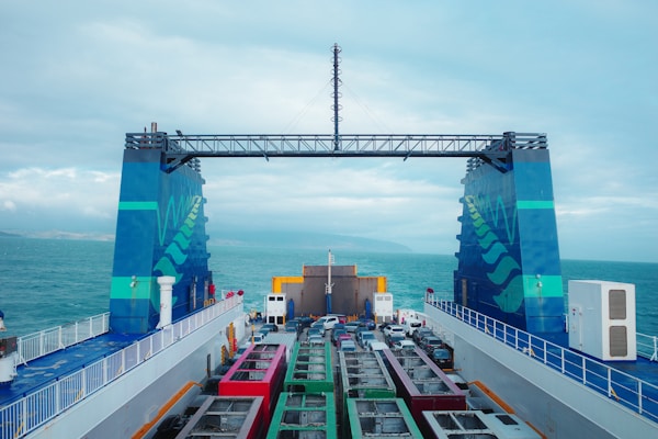A ferry deck filled with various vehicles, including colorful buses and cars, is seen with the open sea in the background. The large ferry has prominent blue structures on either side, adorned with wave patterns, and an overhead metal framework connecting them.