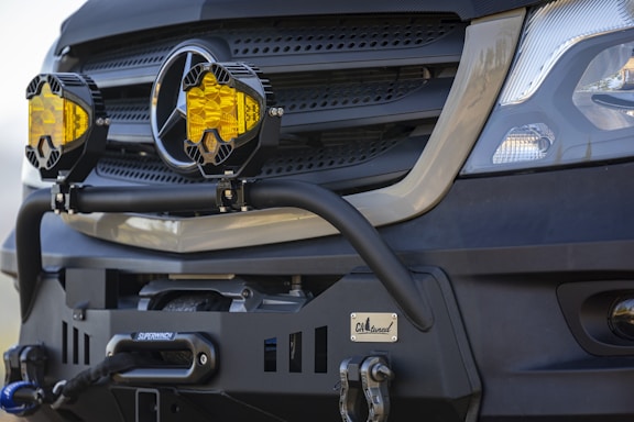 A close-up of a vehicle's front grille, featuring two prominent yellow fog lights mounted. The vehicle has a rugged, off-road bumper with a winch. The branding is visible on the front, and the bumper has a custom badge attached.