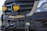 A close-up of a vehicle's front grille, featuring two prominent yellow fog lights mounted. The vehicle has a rugged, off-road bumper with a winch. The branding is visible on the front, and the bumper has a custom badge attached.