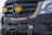 A close-up of a vehicle's front grille, featuring two prominent yellow fog lights mounted. The vehicle has a rugged, off-road bumper with a winch. The branding is visible on the front, and the bumper has a custom badge attached.