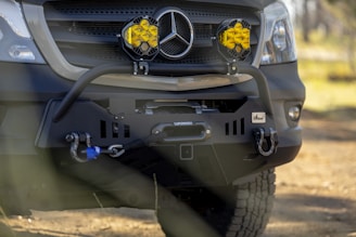 A close-up of a rugged vehicle front bumper featuring a prominent grille with a distinctive emblem. Two round, yellow fog lights are mounted on either side. The bumper has a winch setup with a visible cable, along with heavy-duty hooks.