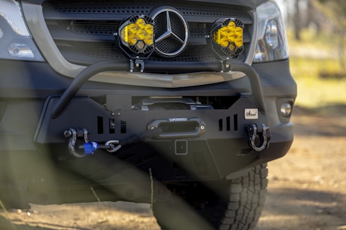 A close-up of a rugged vehicle front bumper featuring a prominent grille with a distinctive emblem. Two round, yellow fog lights are mounted on either side. The bumper has a winch setup with a visible cable, along with heavy-duty hooks.