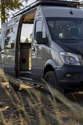 A modern camper van with its side door open, showing a glimpse of the interior. The van is parked in an outdoor setting with a few trees in the background and some dry grass in the foreground. The vehicle has off-road tires and a roof rack, suggesting it's equipped for travel and adventure.