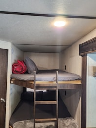 A cozy bunk bed arrangement with a wooden ladder leading to the top bunk, which has a rolled-up red blanket and grey pillows on a grey mattress. The lower bunk is also fitted with dark bedding. The room is softly lit with warm overhead lighting and has plain cream-colored walls.