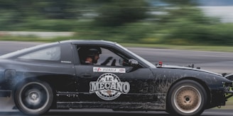 A black sports car is captured in motion on a race track, with a 'Le Mecano' logo prominently displayed on the side. The driver is wearing a helmet, and tire smoke is visible, indicating high speed.