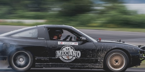 A black sports car is captured in motion on a race track, with a 'Le Mecano' logo prominently displayed on the side. The driver is wearing a helmet, and tire smoke is visible, indicating high speed.