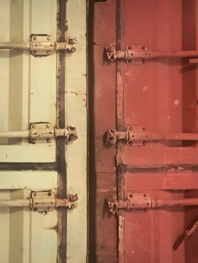 The image features metal doors with bolts and hinges, divided into two sections with distinct colors. On the left, the door is a light cream or pale green shade, while the right side is a reddish-brown. The metal fixtures show signs of wear, giving the doors a rustic or industrial look.