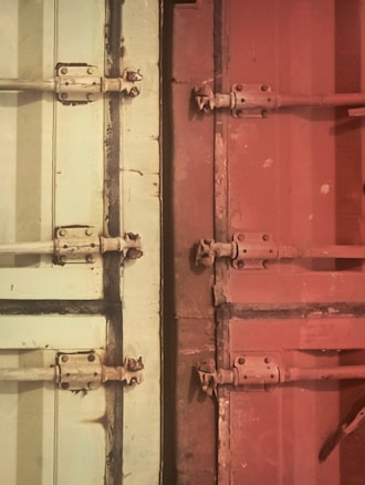 The image features metal doors with bolts and hinges, divided into two sections with distinct colors. On the left, the door is a light cream or pale green shade, while the right side is a reddish-brown. The metal fixtures show signs of wear, giving the doors a rustic or industrial look.