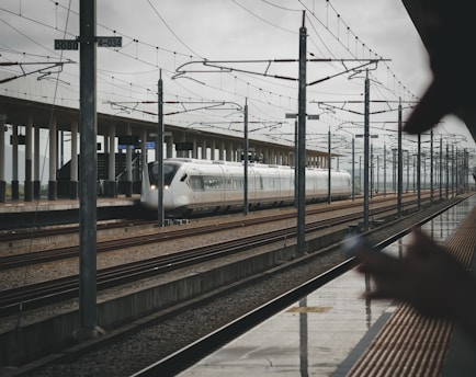 A sleek, modern high-speed train is on railway tracks at a station. Overhead wires and poles stretch along the length of the platform. In the foreground, there is a person holding a smartphone, slightly blurred, suggesting movement or focus on the distant train. The station platform is expansive, with a minimal, contemporary design.