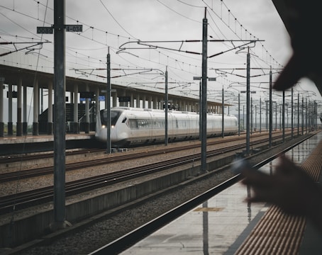 A sleek, modern high-speed train is on railway tracks at a station. Overhead wires and poles stretch along the length of the platform. In the foreground, there is a person holding a smartphone, slightly blurred, suggesting movement or focus on the distant train. The station platform is expansive, with a minimal, contemporary design.