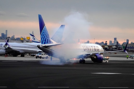 An aircraft with the United Airlines logo is depicted on a runway with smoke or mist around its landing gear area. Several other planes and airport vehicles are visible in the background, along with some airport buildings and structures against a backdrop of an evening or early morning sky.