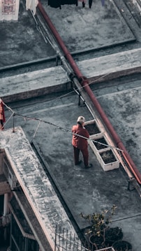 An elderly person wearing a red outfit is standing on a gray rooftop near two rectangular planters. The sky is overcast, and there are pipes and some hanging clothes visible in the background. The rooftop has a rough, worn texture.
