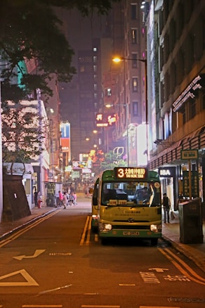 A vibrant urban street scene at night with a minibus parked on the road displaying the route number '3' and destination 'TAI KOK TSUI'. Neon lights illuminate the area, with various shop signs and street lamps casting a warm glow. People can be seen walking along the sidewalk, contributing to the lively city atmosphere.