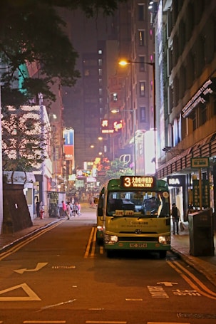 A vibrant urban street scene at night with a minibus parked on the road displaying the route number '3' and destination 'TAI KOK TSUI'. Neon lights illuminate the area, with various shop signs and street lamps casting a warm glow. People can be seen walking along the sidewalk, contributing to the lively city atmosphere.
