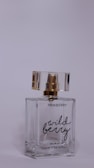 A perfume bottle labeled 'Pinkberry Wild Berry' sits with a transparent body and a gold and clear cap. The bottle is made of glass, displaying a minimalist and elegant design.