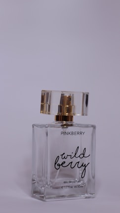 A perfume bottle labeled 'Pinkberry Wild Berry' sits with a transparent body and a gold and clear cap. The bottle is made of glass, displaying a minimalist and elegant design.
