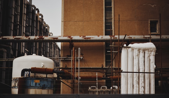 An industrial scene featuring various interconnected machinery and pipes mounted on a building. The image shows a large cylindrical tank with rusted sections and insulated piping. Several vertical and horizontal metal pipes are visible, some of which show signs of rust. The background consists of an old, weathered wall with small windows, and a piece of industrial equipment is wrapped with white insulation material.