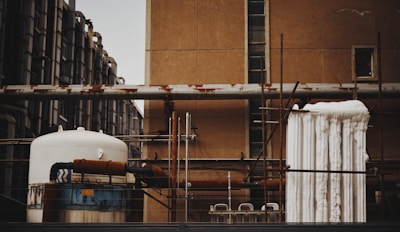 An industrial scene featuring various interconnected machinery and pipes mounted on a building. The image shows a large cylindrical tank with rusted sections and insulated piping. Several vertical and horizontal metal pipes are visible, some of which show signs of rust. The background consists of an old, weathered wall with small windows, and a piece of industrial equipment is wrapped with white insulation material.