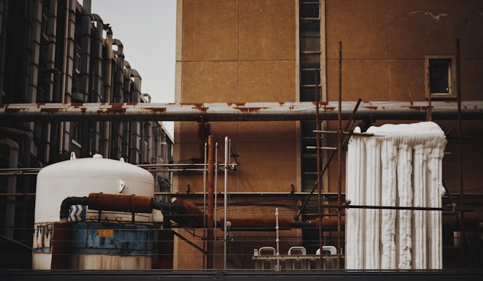An industrial scene featuring various interconnected machinery and pipes mounted on a building. The image shows a large cylindrical tank with rusted sections and insulated piping. Several vertical and horizontal metal pipes are visible, some of which show signs of rust. The background consists of an old, weathered wall with small windows, and a piece of industrial equipment is wrapped with white insulation material.