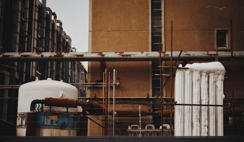 An industrial scene featuring various interconnected machinery and pipes mounted on a building. The image shows a large cylindrical tank with rusted sections and insulated piping. Several vertical and horizontal metal pipes are visible, some of which show signs of rust. The background consists of an old, weathered wall with small windows, and a piece of industrial equipment is wrapped with white insulation material.