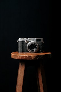 A vintage-style camera is placed centrally on a wooden stool against a dark background, creating a minimalist and classic aesthetic.