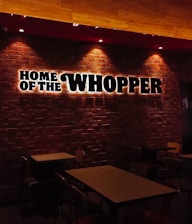 Dimly lit restaurant interior with a brick wall featuring illuminated signage saying 'Home of the Whopper.' The image shows empty tables and chairs under warm lighting, creating a cozy atmosphere.