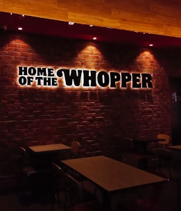 Dimly lit restaurant interior with a brick wall featuring illuminated signage saying 'Home of the Whopper.' The image shows empty tables and chairs under warm lighting, creating a cozy atmosphere.