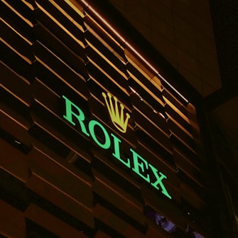An illuminated sign with the brand name 'ROLEX' in green neon lights and a golden crown logo above, set against a dark, slatted background.