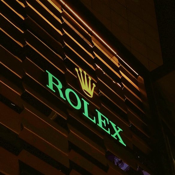 An illuminated sign with the brand name 'ROLEX' in green neon lights and a golden crown logo above, set against a dark, slatted background.