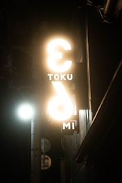 A dimly lit street scene featuring a glowing vertical sign with partially visible letters, including 'TOKU' and 'IMI'. The sign has a modern design with prominent light emitting from it, casting a warm glow. A streetlight is visible in the background, illuminating the surrounding area slightly.