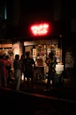 A small group of people stands outside a dimly lit storefront at night. A bright neon sign glows red above the entrance, while several posters and signs are displayed in the window and around the shop. The atmosphere suggests a late-night hangout spot or a small bar or restaurant.