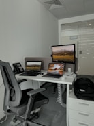 A modern office setup featuring a white desk with a laptop, a monitor displaying a landscape, a black office chair, and a telephone. The space also includes a backpack on a set of drawers and a glass door in the background.