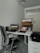 A modern office setup featuring a white desk with a laptop, a monitor displaying a landscape, a black office chair, and a telephone. The space also includes a backpack on a set of drawers and a glass door in the background.