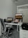 A modern office setup featuring a white desk with a laptop, a monitor displaying a landscape, a black office chair, and a telephone. The space also includes a backpack on a set of drawers and a glass door in the background.