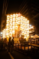 A vibrant festival scene at night, featuring a large arrangement of illuminated paper lanterns with traditional symbols and characters. The lanterns are arranged in a grid and glow brightly against the dark surroundings. People are gathered around, some observing and some engaged in activities, creating a lively atmosphere. Traditional wooden structures and hangings add to the cultural setting.