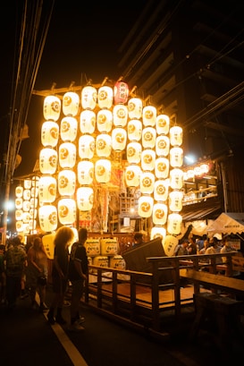 A vibrant festival scene at night, featuring a large arrangement of illuminated paper lanterns with traditional symbols and characters. The lanterns are arranged in a grid and glow brightly against the dark surroundings. People are gathered around, some observing and some engaged in activities, creating a lively atmosphere. Traditional wooden structures and hangings add to the cultural setting.